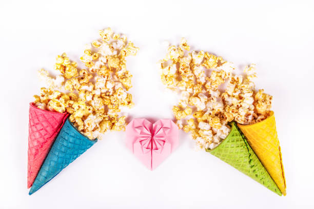 Sweet caramel popcorn and paper heart. Copy space. Origami heart of pink paper. Bright festive sweets. Colored waffle cones stock photo