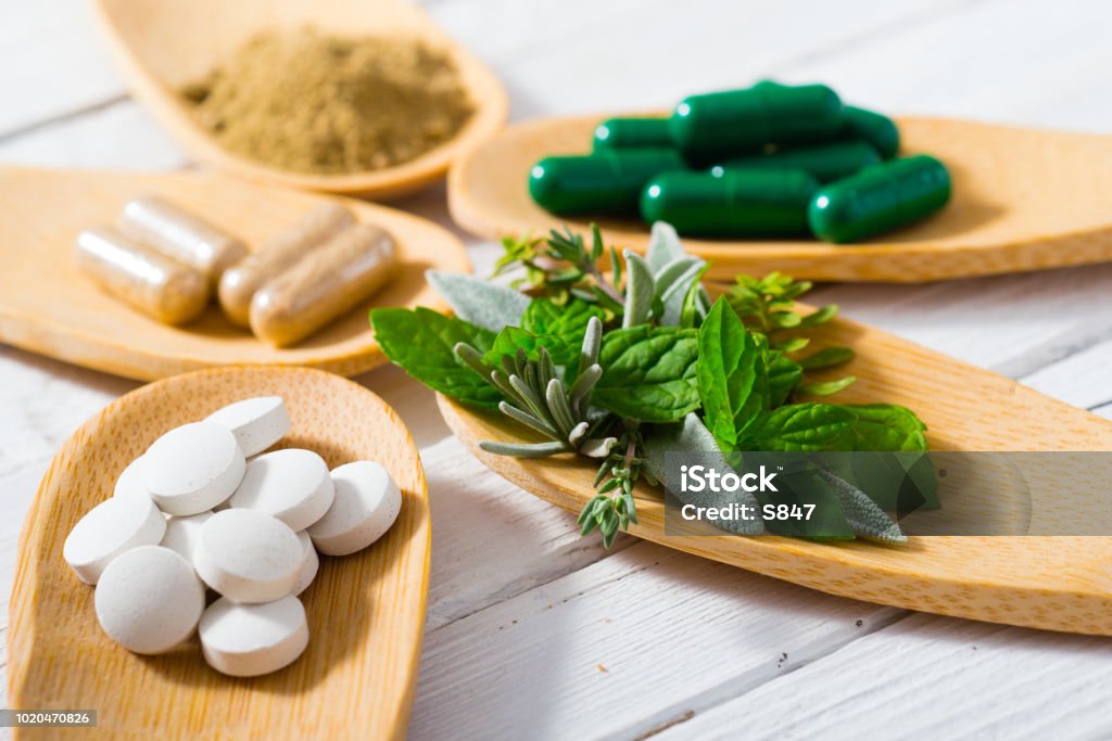 Medicines herbal leaves, ground herb powder and medicament pills on bamboo spoons, white wooden table Nutritional Supplement Stock Photo