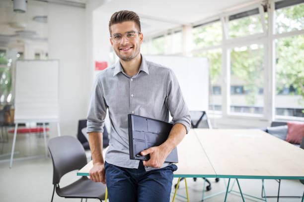 Confident businessman standing by conference table Portrait of handsome young businessman standing by conference table in new office. Confident man with file looking at camera and smiling. one young man only stock pictures, royalty-free photos & images