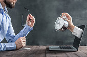 Businessman is sitting at the desk and is looking at the hand with clock coming out of the laptop