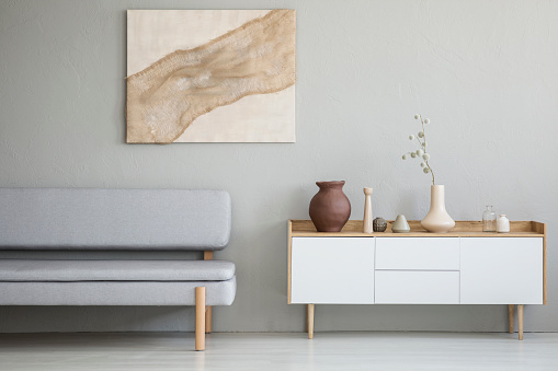 Real photo of a simple living room interior with a natural painting on the wall and gray sofa next to a wooden cupboard