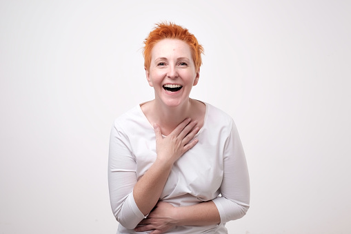 Close up portrait of a beautiful mid adult woman with red hair laughing standing on gray background. She is impressed and out of mind
