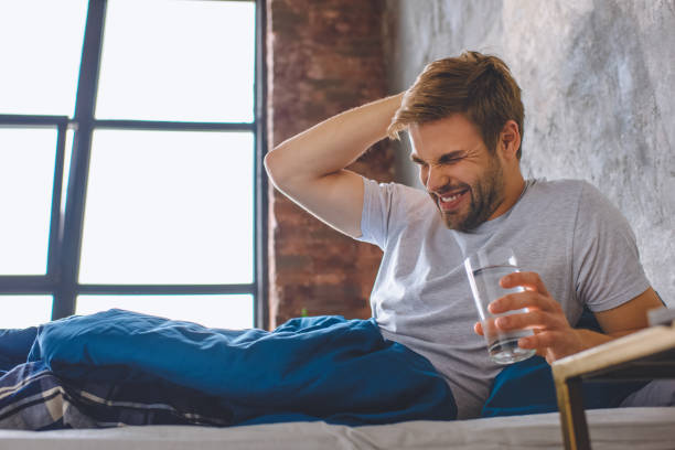 young man with headache holding glass of water in bed at home young man with headache holding glass of water in bed at home hangover stock pictures, royalty-free photos & images