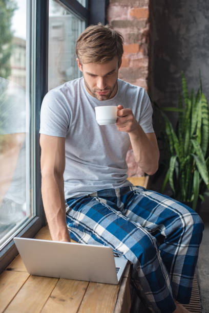 istock young man with coffee cup using laptop on windowsill at home 1020561698