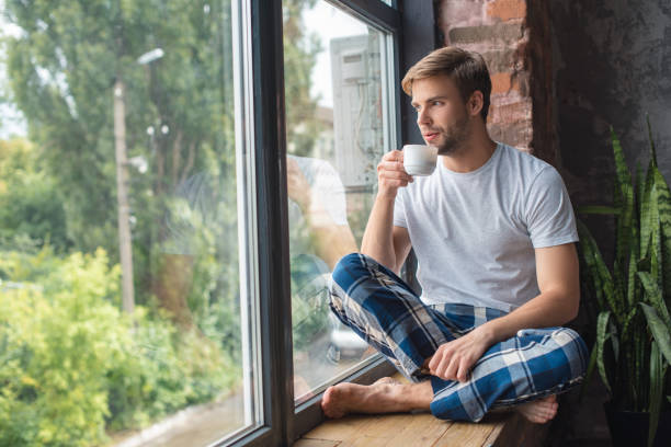 まじめな若者窓辺に座って、朝の時間を自宅の中にコーヒーを飲む - window light window sill home interior ストックフォトと画像