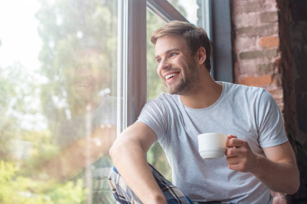 istock smiling man looking in windows and sitting with cup of coffee on windowsill 1020563130