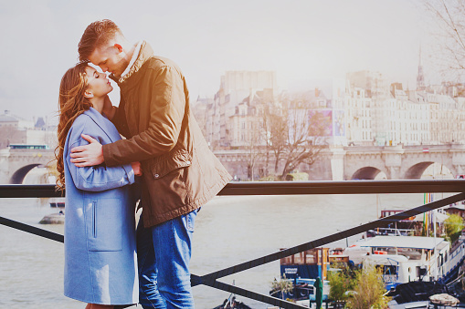 love couple kissing on the street in Paris, romantic moment in honeymoon, happy affectionate young man and woman standing on bridge over Seine river