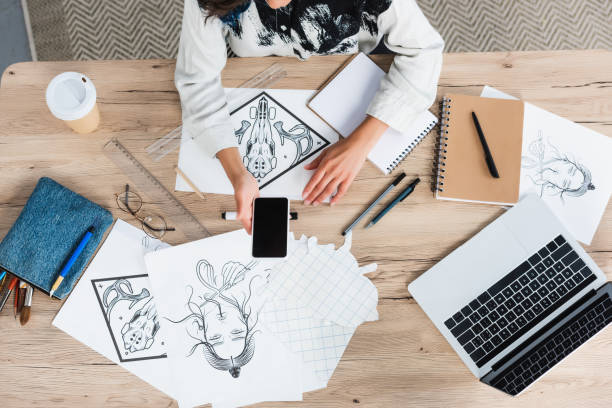 istock cropped image of female designer holding smartphone with blank screen at working table with paintings and textbooks 1020616570