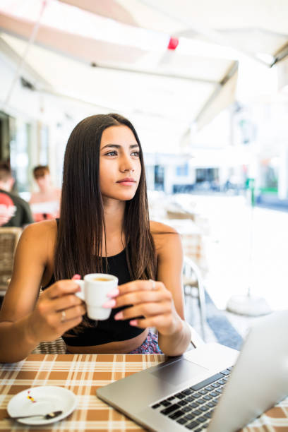 istock Young female student drink coffee while keyboarding on her laptop computer while sitting in cafe in the fresh air in summer day. Pretty latin woman connecting to wireless via net-book during morning breakfast 1020673918