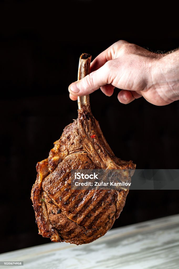 Um grande pedaço de assado de carne de carneiro, carne de porco, vitela ou carne nos ossos cozidos em uma fritadeira de ar, nas mãos do chefe do chefe, uma imagem de fundo em um fundo preto. - Foto de stock de Acima royalty-free