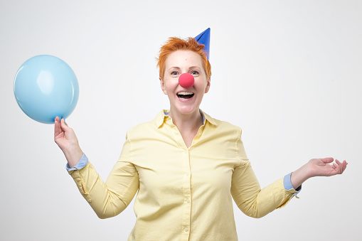 Pretty mature happy woman with colorful balloon and red clown nose on blue background. She celebrates her birthday or congratulates her friend