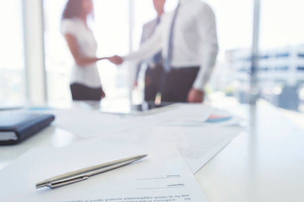 Business woman and business man shaking hands with a contract. Business woman and business man shaking hands with a contract. There is a pen on the contract document. Focus is on the foreground with the three people in the office out of focus in the background. They are wearing formal corporate business clothes. contract stock pictures, royalty-free photos & images