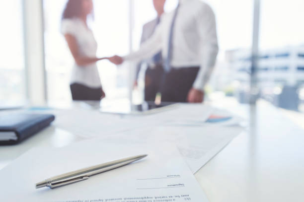 Business woman and business man shaking hands with a contract. There is a pen on the contract document. Focus is on the foreground with the three people in the office out of focus in the background. They are wearing formal corporate business clothes.