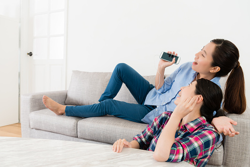 cheerful beauty girl lying down on sofa couch and using mobile smartphone connect headphone listening music singing with friend.