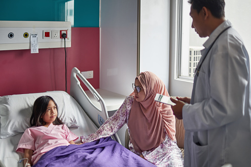 Mature doctor and woman looking at girl on bed in ward. Patient is with mother and healthcare worker. They are at hospital.