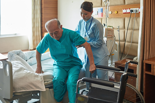 Nurse assisting senior patient to sit on bed. Female healthcare worker taking care of man in hospital. Image is representing assisted living.