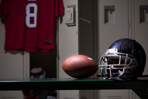 Various sports equipment on bench inside high school or college gymnasium locker room.   Items include: football helmet, ball, pads, jersey.