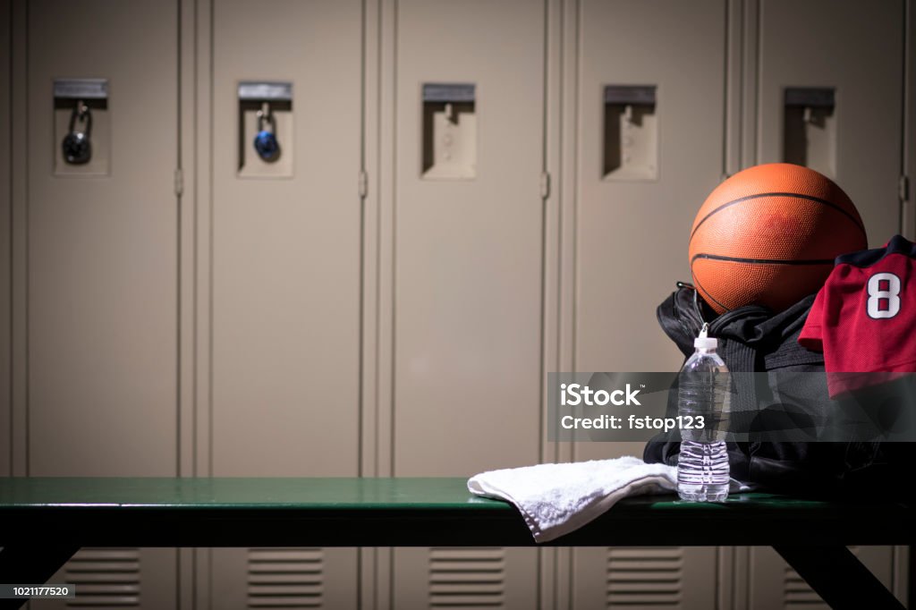 Basketball sports equipment in school gymnasium locker room. Various sports equipment on bench inside high school or college gymnasium locker room.   Items include: basketball, jersey, water, towel, gym bag.. Basketball - Sport Stock Photo