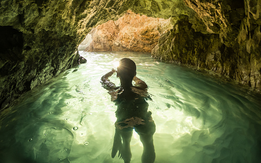 Young woman swimming in water cave inside the mountain toward exit with light