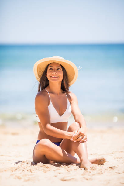 istock Young latin attractive in white bikini and straw hat posing while sitting on the beach 1021199762