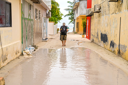 Small street in the Maafushi island in Maldives flooded with rain water, a senior man with backpack, rear view, walking to the sea in distance.