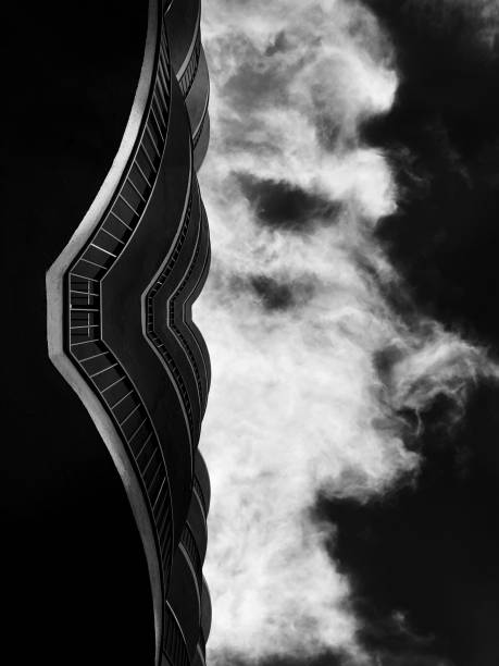 A black and white photograph showcases a towering building that is prominently emitting wisps of smoke from its structure into the sky above