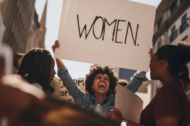 African woman holding a banner and laughing during women march. Group of female demonstrating outdoors with placards.