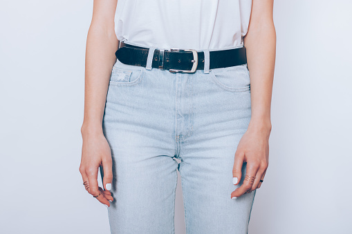 Slender young woman wearing blue high waist mom's jeans with belt and plain t-shirt standing over white background, close-up.
