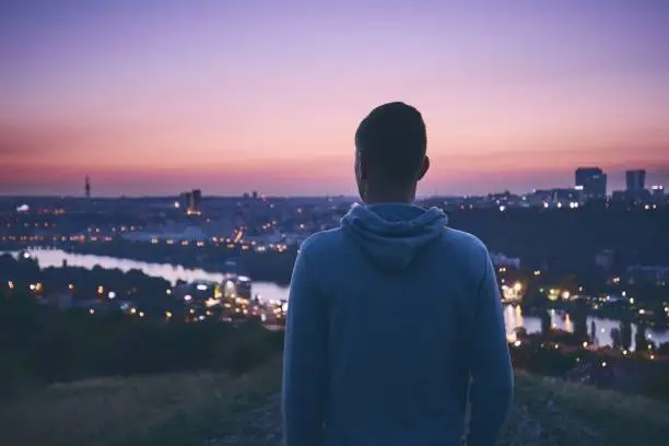 Silhouette of the pensive young man waiting for sunrise on the hill against urban skyline. Prague, Czech Republic.