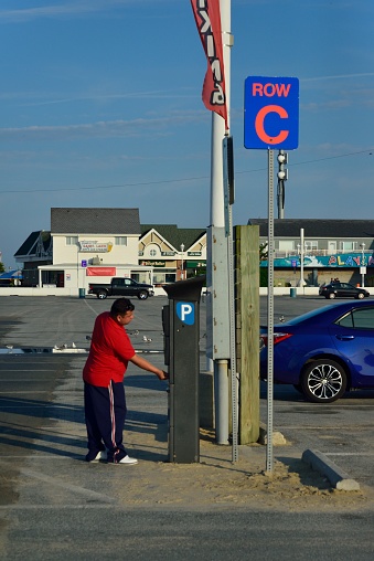 A hot summer Sunday morning  at the Ocean City Inlet where tourist have started to gather, a lone hispanic man uses the automated parking station to register his car and retrieve his parking pass with the boardwalk cityscape in the background   Photo by Bob Balestri, dba Joesboy
