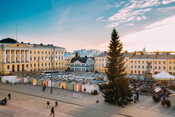 Photo of Helsinki, Finland. People Walking On Christmas Xmas Market With Christmas Tree On Senate Square In Background Of Government Palace At Winter Sunny Day
