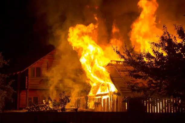 Photo of Burning wooden house at night. Bright orange flames and dense smoke from under the tiled roof on dark sky, trees silhouettes and residential neighbor cottage background. Disaster and danger concept.