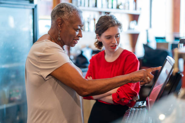 aktywny starszy, 77-letni, african-american bizneswoman, właściciel firmy, nauczanie nowego pracownika, 18-letni kaukaski białej dziewczyny, jak korzystać z skomputeryzowanej kasy w małej lokalnej restauracji. - 18 19 years happiness cheerful teenager zdjęcia i obrazy z banku zdjęć