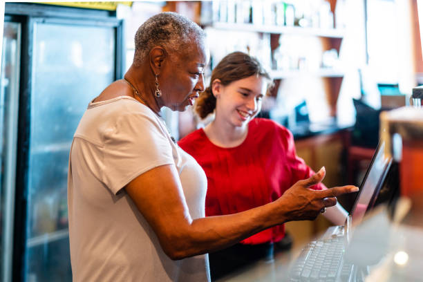 the active senior, 77-years-old, african-american businesswoman, business owner, teaching the new employee, the 18-years-old caucasian white girl, how to use the computerized cash register in the small local restaurant. - independence lifestyles smiling years imagens e fotografias de stock