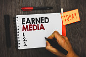 Text sign showing Earned Media. Conceptual photo Publicity gained through promotional efforts by multimedia Man holding marker notebook clothespin hold reminder cup markers wood table.