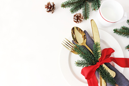 Traditional Christmas table place setting. Golden cutlery, linen napkin, spruce branches and cup of milk. Pine cone and red ribbon decoration. Holidays background, flat lay, top view with copy space.