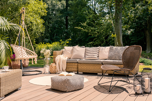Pouf and rattan chair on wooden patio with settee in the garden during summer. Real photo