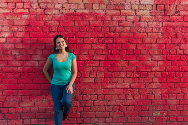 A millennial eurasian woman is casually leaning against a red brick wall on a sunny day