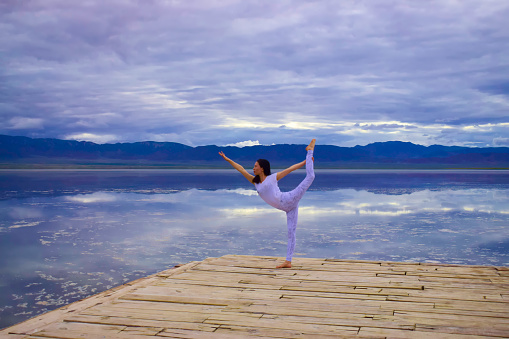 Landscape. A yoga dancer practices on the dock in front  of a salt lake, clouds, mountains and reflections as back ground. Created in Qinghai, China. 07/05/2018.