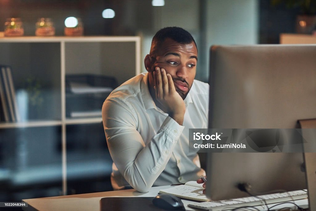 Staring at the screen but nothing's going in Shot of a young businessman looking bored while working at his desk during late night at work Boredom Stock Photo