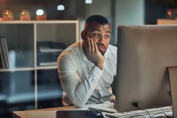 Shot of a young businessman looking bored while working at his desk during late night at work