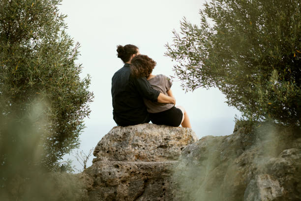 Rear view of hugging couple sitting on natural rocks among olive trees
