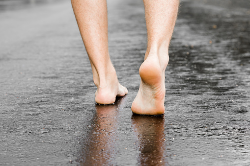 Young man's barefoot walking on the wet, dark black asphalt after warm rain. Cloudy day in summer. Back view.