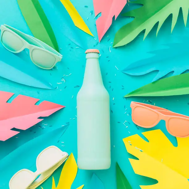 Photo of Painted in blue beer or cocktail bottle lies among sunglasses and palm tropical leaves