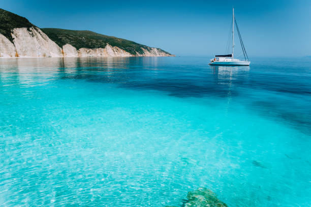 einsamen weißen segeln katamaran boot drift auf ruhiger see oberfläche. reinen seichten azurblauen bucht wasser an einem schönen strand. malerische landschaft der felsigen küste im hintergrund - solitude remote sailboat horizontal stock-fotos und bilder