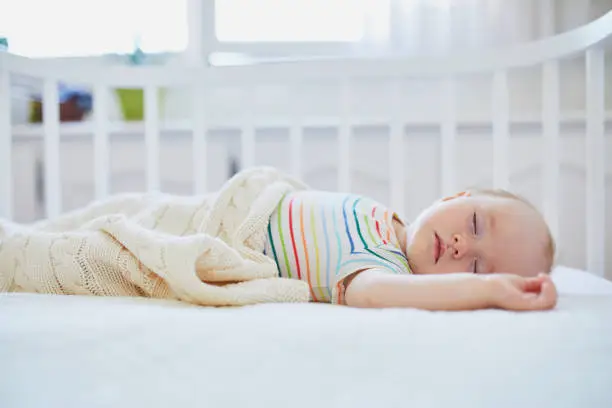 Adorable baby girl sleeping in co-sleeper crib attached to parents' bed. Little child having a day nap in cot. Infant kid in sunny nursery