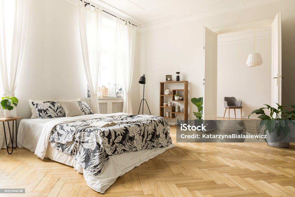 Stylish apartment interior with white walls and herringbone wooden floor. A view from a bedroom with a big bed to another room with an armchair. Real photo. Bedroom Stock Photo