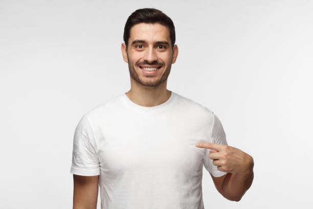 istock Young man pointing with index finger at blank white t-shirt with empty space for your advertising text or image, standing isolated on gray background 1022174450
