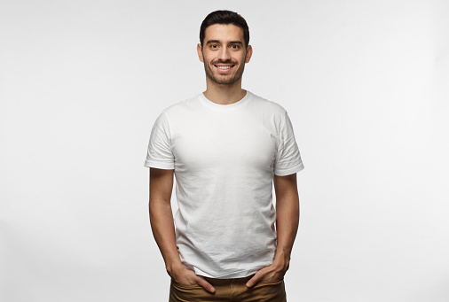 Young european man standing with hands in pockets, wearing blank white tshirt with copy space for your logo or text, isolated on grey background