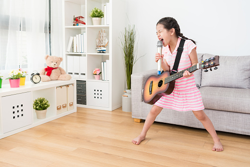 Little Asian girl sinks herself into singing with microphone mike and playing guitar. She gives a professional performance in the living room. She enjoying the music.